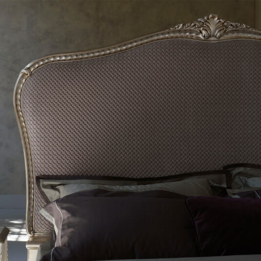 A close-up of the High End Classic Style Upholstered Bed reveals its intricate wooden carvings at the top. The bed is adorned with dark-colored linens and pillows, while soft light casts gentle shadows on the textured headboard.