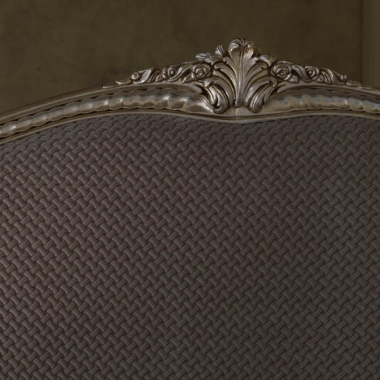 The High End Classic Style Upholstered Bed boasts an ornate headboard featuring intricate carvings and a woven pattern in a dark finish. The top edge showcases detailed, elegant scrollwork, infusing luxury into the classic style design. The muted background accentuates the bed's exceptional craftsmanship.