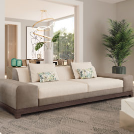 Create a calm space: A modern, spacious living room featuring a High-End Deluxe Three-Seater Sofa in beige and brown, adorned with patterned cushions. A tall green plant stands to the right, and in the background, a dining area with a contemporary chandelier and minimalist artwork is visible through a wide doorway.