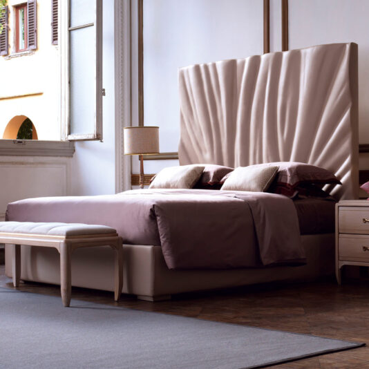 A modern bedroom showcasing the High End Leather Bed With Tall Headboard, characterized by its textured beige headboard and brown bedding. Two nightstands with lamps frame the bed, which also features a cushioned bench at its foot. Natural light streams in through an open window pane.