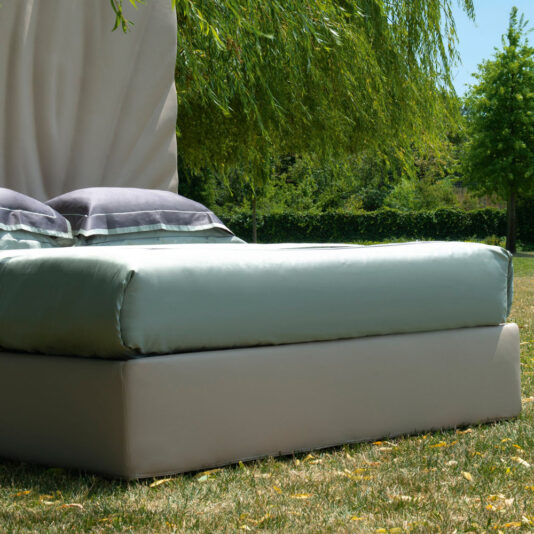 High End Leather Bed With Tall Headboard A High End Leather Bed With Tall Headboard, adorned with white sheets and two gray pillows, is situated on a grassy area beneath a leafy green tree. The surrounding landscape boasts lush greenery with trees in the background on a sunny day.