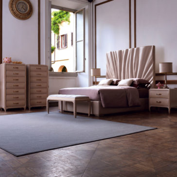 High End Leather Bed With Tall Headboard Experience comfort and style in this spacious, elegant bedroom featuring our High End Leather Bed With Tall Headboard. The luxurious bed boasts a tall, wavy upholstered headboard that makes a statement. Complementing the bed are two matching nightstands, a dresser, and a tall chest of drawers. A bench positioned at the foot of the bed offers additional seating or storage space. The room's open window lets in natural light and offers views of lush greenery outside, while the wooden floor adds warmth to this inviting space.