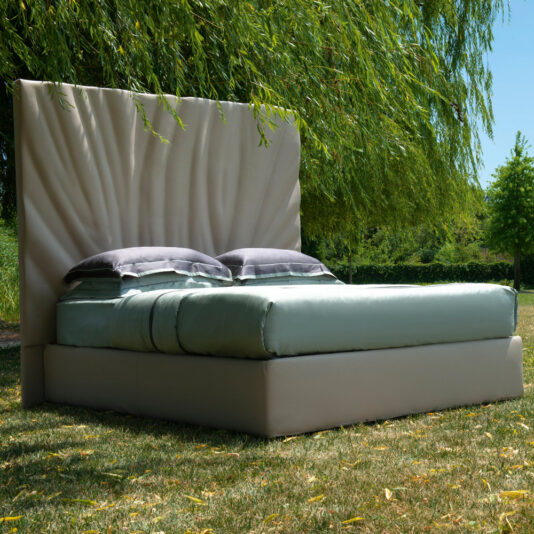 High End Leather Bed With Tall Headboard A High End Leather Bed With Tall Headboard is set outdoors on a grassy area beneath a tree with lush, drooping branches. The bed is neatly made with pale blue bedding and two gray pillows, and the background features other trees and a clear, blue sky.