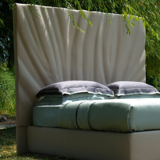 High End Leather Bed With Tall Headboard The High End Leather Bed With Tall Headboard, featuring a textured beige headboard, is placed under green, leafy branches. Made with light-green sheets and two stacked pillows with gray covers, the setup creates a cozy and serene setting in the lush, green surroundings.