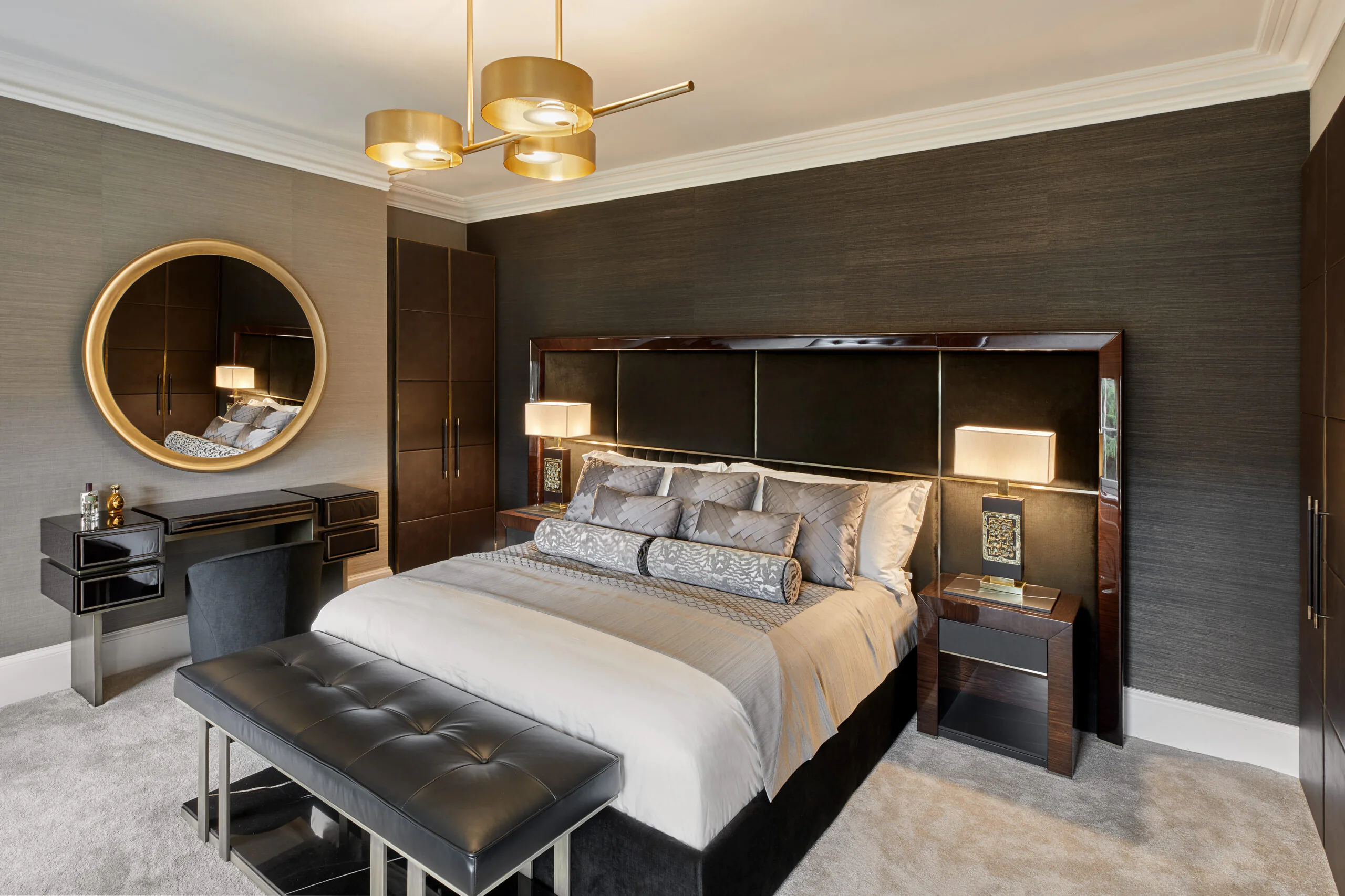 A modern bedroom features a luxurious bed with plush gray and white bedding. Two bedside tables with lamps flank the bed. A round mirror hangs above a sleek vanity with drawers. A bench sits at the foot of the bed, and a chandelier hangs from the ceiling.