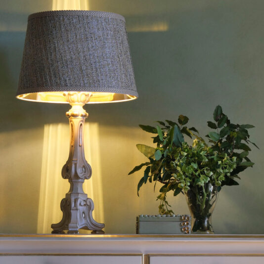 The Large Carved Italian Table Lamp, adorned with a textured grey lampshade, casts a warm light against a green wall. Beside the lamp, there is a lush arrangement of leafy green plants in a clear glass vase. The setup is placed on a sleek, white console table.