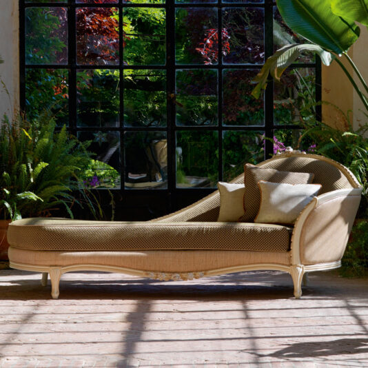 A Large Classic Louis Style Chaise Longue is placed on a sunlit wooden floor in front of a spacious, black-framed window. The space is adorned with various green plants and trees, creating a serene and inviting atmosphere.