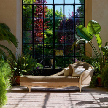 Large Classic Louis Style Chaise Longue A Large Classic Louis Style Chaise Longue with cushions is set in a sunlit room with expansive grid-like windows that overlook a lush garden. The lounge is surrounded by large potted plants, creating a peaceful and inviting atmosphere.