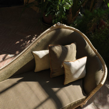 Large Classic Louis Style Chaise Longue The Large Classic Louis Style Chaise Longue, featuring a textured beige pattern, is complemented by two square cushions—one in beige and the other in light brown. Situated in a bright area bathed in natural sunlight and shadows, it is encircled by potted plants with lush green foliage.