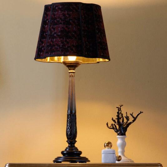 Large Luxury Carved Table Lamp A Large Luxury Carved Table Lamp with an ornate, dark base and a textured, dark red lampshade is illuminated. Beside it is a white ceramic decorative piece resembling a tree and a small white container with a gold lid, all set against a soft yellow wall.