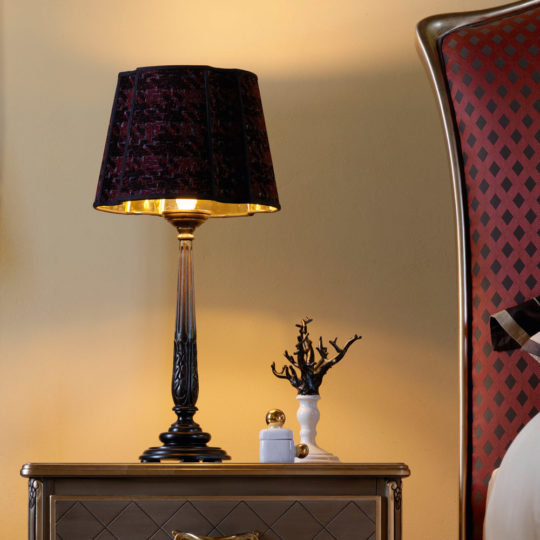 A Large Luxury Carved Table Lamp with a black base and a dark patterned shade is placed on a wooden bedside table. Beside it, there’s a small figurine and an abstract, tree-like decor piece. The backdrop features a red and black-patterned headboard against a muted yellow wall.
