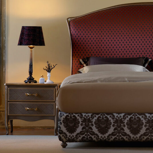 Large Luxury Carved Table Lamp A bedroom showcases an elegant bed with a headboard adorned in a sophisticated red and black pattern, complemented by a beige bedspread. Next to it, a classy bedside table prominently displays the Large Luxury Carved Table Lamp, featuring an ornate dark shade. A small white ornament adds an extra touch of refinement to the setting. The room is gently illuminated with warm tones, creating a cozy ambiance.