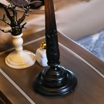 Large Luxury Carved Table Lamp A close-up of a bedside table showcasing the Large Luxury Carved Table Lamp with its detailed base. To the left, there's a white ceramic ring holder shaped like a tree and a small white jar with a golden lid. The background captures part of the bed and pillow adorned with blue patterns.