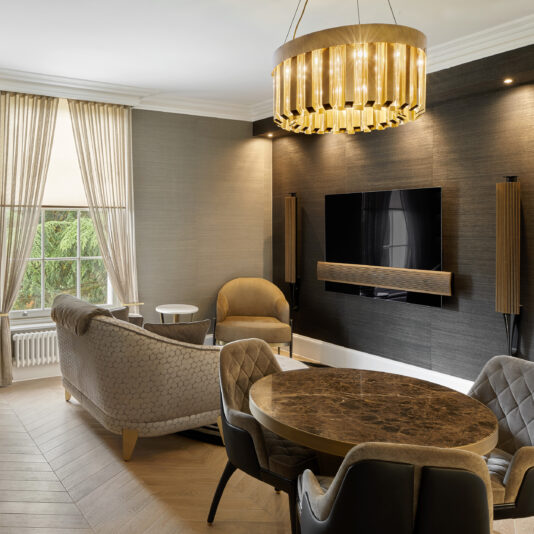 A modern living room featuring a black accent wall with a mounted flat-screen TV and speakers. A marble dining table with upholstered chairs sits under the Luxurious Round Designer Chandelier. Light gray sofas and a large window with beige curtains complete the elegant design.