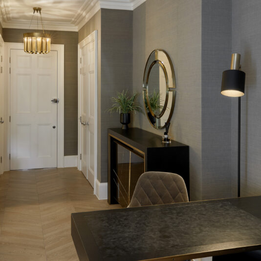 A modern office space features a sleek desk and gray upholstered chair in the foreground. A circular mirror, potted plant, and black lamp adorn a console table against a textured gray wall. The elegant touch of the Luxurious Round Designer Chandelier complements the chevron-patterned wooden flooring and white door.