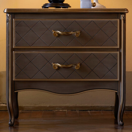 Luxury Italian Bedside Cabinet The Luxury Italian Bedside Cabinet is a vintage wooden nightstand featuring two drawers adorned with a diamond pattern design. Each drawer comes with ornate gold handles showcasing a scroll design. Set against a beige wall, this nightstand exudes timeless elegance with its polished, slightly curved base.