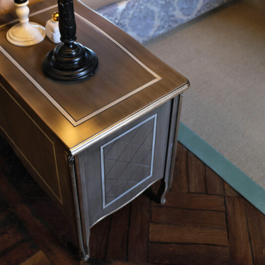 Luxury Italian Bedside Cabinet The Luxury Italian Bedside Cabinet, with its dark wooden top, decorative inlay, and ornate lamp, is gracefully positioned on a parquet floor next to a finely textured rug. The cabinet boasts carved details and polished surfaces, featuring intricate patterns on its front panel.
