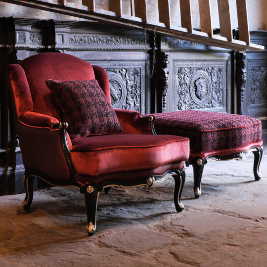 The Luxury Louis Style Armchair and Footstool, upholstered in opulent red velvet, stands on a stone floor against a decorative, dark wood-paneled wall. The armchair features elegant black and gold accents, with a patterned red pillow resting on it to enhance the luxurious setting.