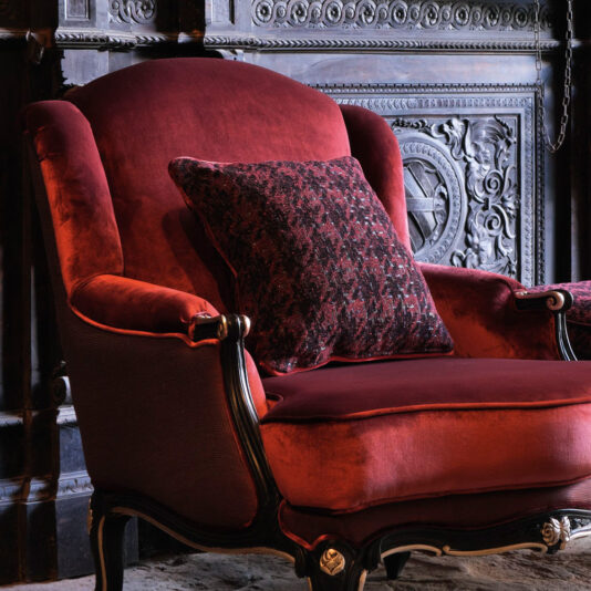 A plush, Luxury Louis Style Armchair with ornate dark wooden legs and armrests sits against a decorative, intricately carved wooden wall. A matching patterned cushion rests against the back of the chair, adding a touch of elegance to the cozy setting. The ensemble is completed with a luxurious footstool from the same collection.