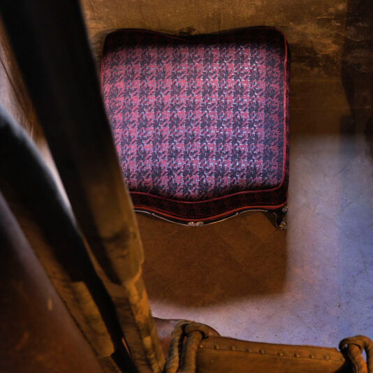 An overhead view of a red upholstered seat featuring an intricate purple and black pattern is positioned next to a worn, brown leather armchair on a tiled floor. The lighting casts shadows, creating a moody ambiance reminiscent of the Luxury Louis Style Armchair And Footstool setting.