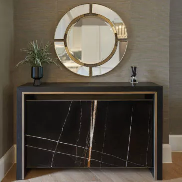 Luxury Marble Radiator Cover Console The Luxury Marble Radiator Cover Console, with its sleek black marble finish, stands proudly against a textured wall. Atop the console sits a black vase filled with lush green foliage alongside an elegant diffuser. Mounted above the console is a large round mirror featuring segmented gold accents, capturing the very essence of luxury.