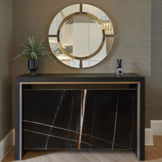 The Luxury Marble Radiator Cover Console, with its sleek black marble finish, stands proudly against a textured wall. Atop the console sits a black vase filled with lush green foliage alongside an elegant diffuser. Mounted above the console is a large round mirror featuring segmented gold accents, capturing the very essence of luxury.