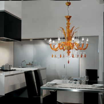 A modern kitchen with a sleek black and white color scheme features the exquisite Luxury Orange Crystal Drop Chandelier hanging above a rectangular dining table set for two, complete with elegant glassware and place settings. The kitchen also boasts contemporary cabinets and appliances.