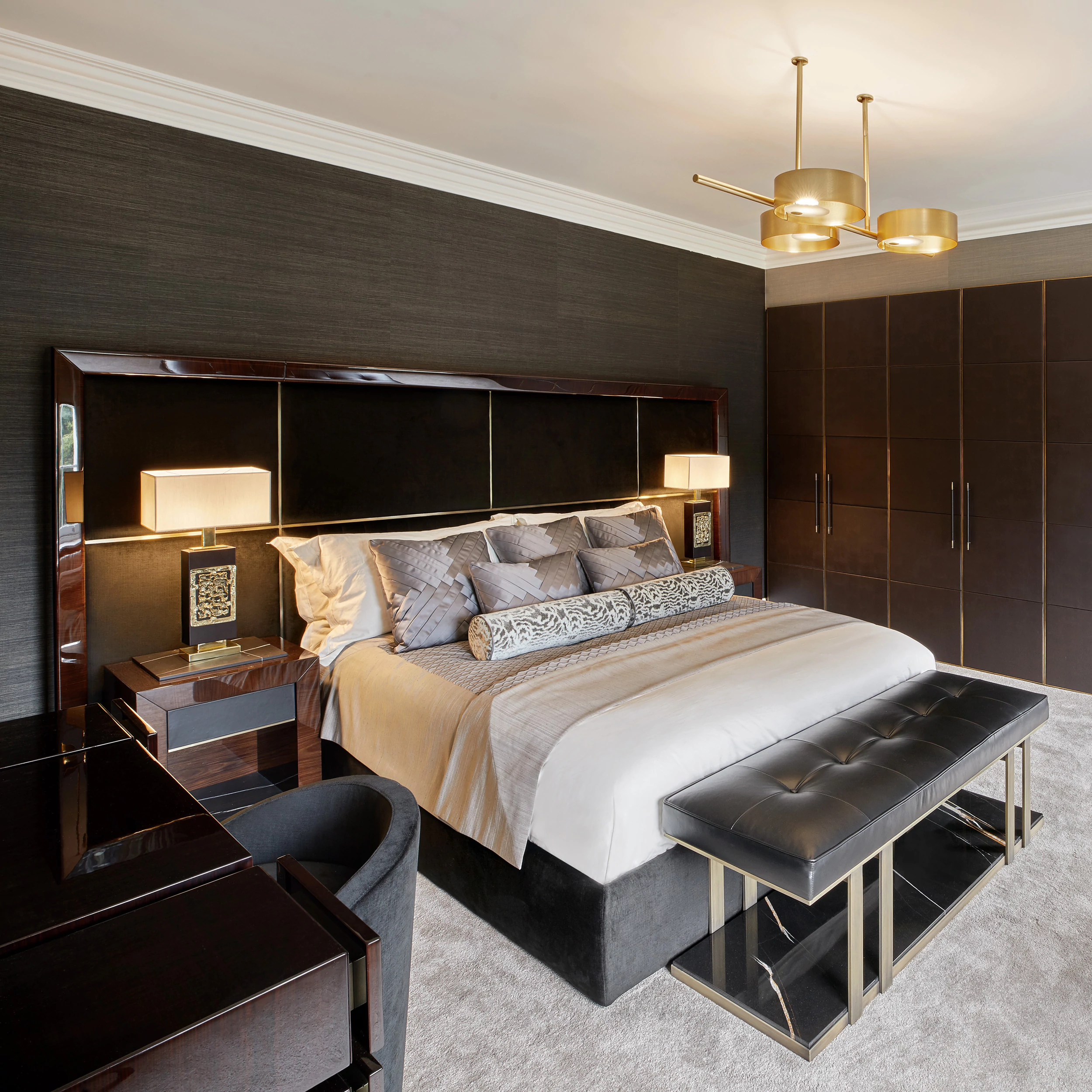 Luxurious bedroom with a modern design. Features a large bed with patterned pillows and a rolled cushion, dark wood headboard, and gold-accented nightstands. A black bench sits at the foot of the bed, and a stylish chandelier hangs overhead.