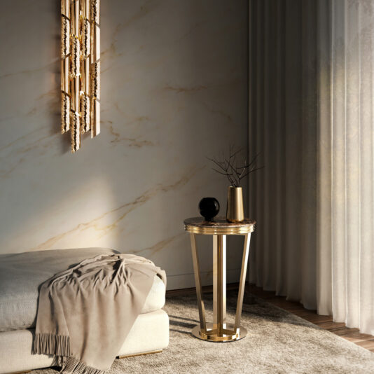 A modern, minimalistic living space featuring a plush beige couch draped with a light gray throw. Beside it sits our **Luxury Round Marble Side Table**, which holds a black vase with bare branches and a black decorative sphere. The background includes marble walls, a gold chandelier, and sheer curtains.
