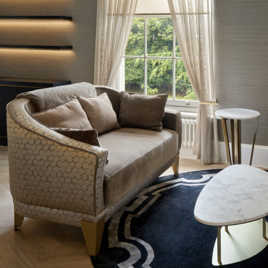 A stylish living room features the Luxury Small Two-Tone Sofa with gold-accented legs, decorated with multiple cushions. Nearby, a modern oval coffee table and a side table complete the look. A large window with sheer curtains overlooks a lush green landscape.