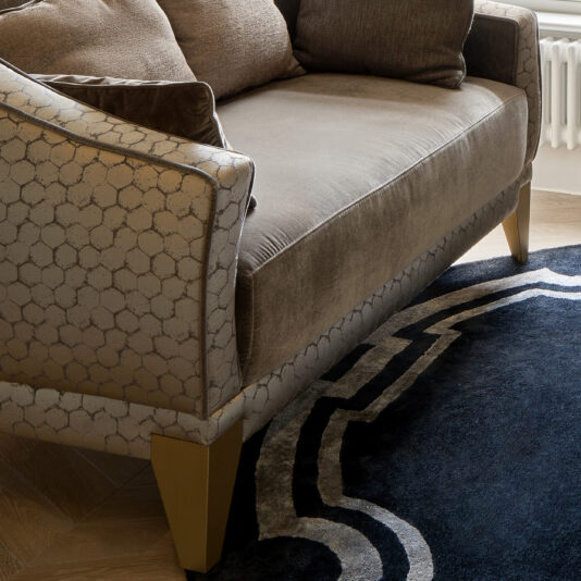 Luxury Small Two-Tone Sofa A close-up view of the Luxury Small Two-Tone Sofa, featuring patterned fabric on the armrests and backrest. It boasts two large square cushions and is situated on a wooden floor next to a plush, dark-colored rug with geometric designs, near a radiator under a window.