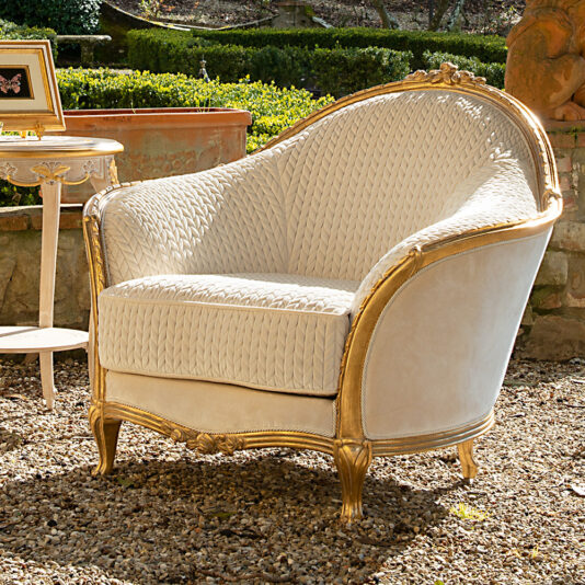 Luxury Venetian Style Armchair A Luxury Venetian Style Armchair, upholstered in elegant beige and gold fabric, featuring intricate wood carvings, is placed outdoors on a gravel surface. A small side table with a framed butterfly picture accompanies the armchair. Greenery and a stone wall provide a scenic backdrop.