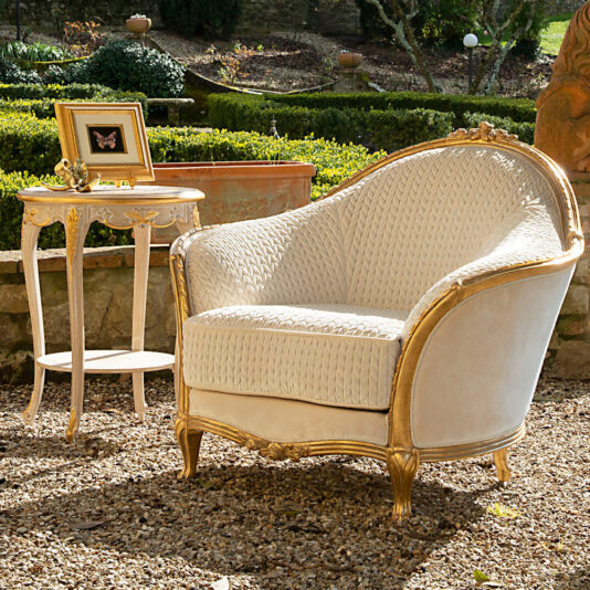 A luxury Venetian style armchair in white and gold sits on a gravel path in a picturesque garden. Nearby, a matching small round side table holds a framed butterfly picture and a decorative gold figurine. The background showcases lush greenery and beautifully landscaped garden features.