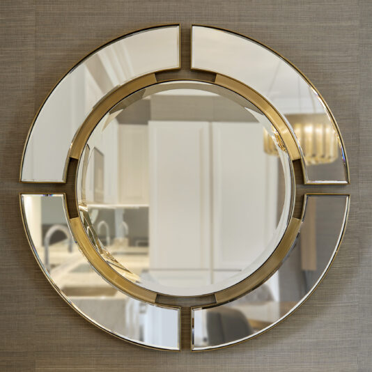 The Modern Designer Round Mirror, featuring a segmented design, is displayed on a textured wall. It reflects the contemporary kitchen interior, showcasing sleek white cabinets, a spacious countertop, and an elegant decorative light fixture.