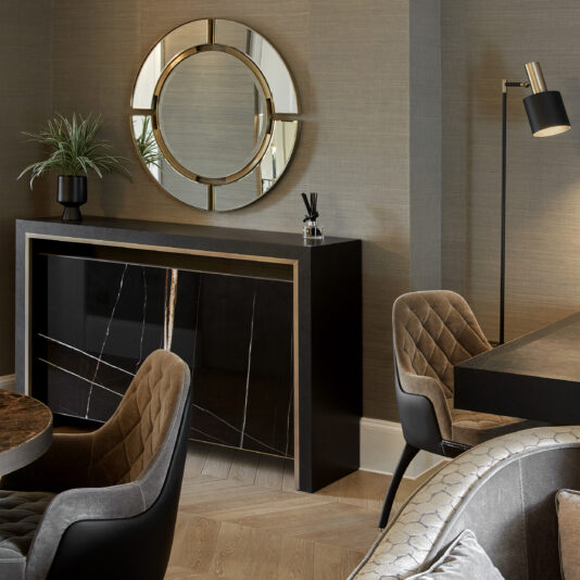 A modern room showcases the Modern Designer Round Mirror above a sleek, black sideboard with gold accents. Quilted grey chairs surround a wooden table, and a tall floor lamp stands nearby. A potted plant and decorative items are displayed on the sideboard, enhancing the modern designer aesthetic.