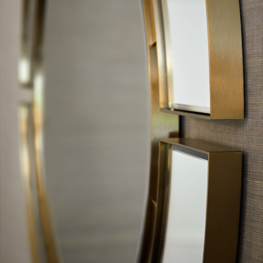 A close-up of the Modern Designer Round Mirror, featuring three vertically aligned round segments with a gold frame. The decorative mirror reflects a blurred background, highlighting its sleek and minimalist design against a textured wall.