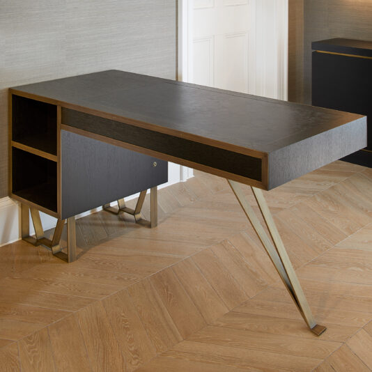 The Modern Industrial Style Oak Veneer Desk boasts a sleek design and a dark oak veneer finish. It features a minimalist appearance with unique angled legs, shelves on the left side, and is set against a backdrop of wooden flooring and light-colored walls.