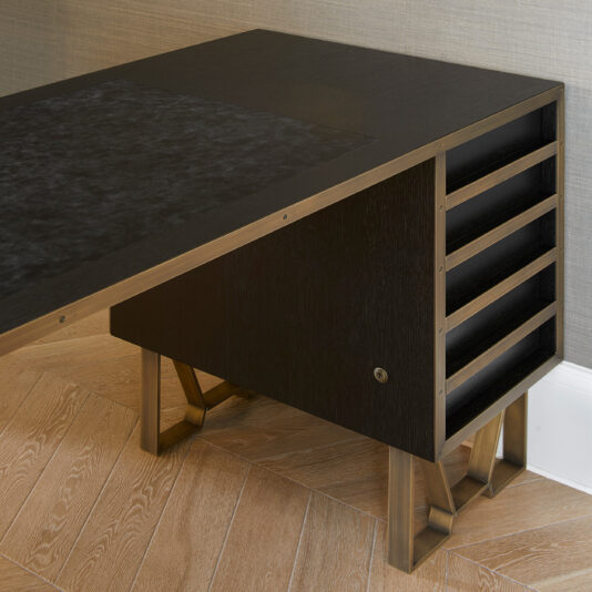 A sleek Modern Industrial Style Oak Veneer Desk with a minimalist design placed against a grey wall. The desk features angled metal legs and a drawer unit with multiple open slots on the right side. The surface has a dark, textured area resembling a desktop mat. The wooden floor enhances its oak veneer finish.
