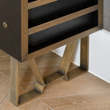 Modern Industrial Style Oak Veneer Desk Close-up of a corner of a modern, wooden cabinet with brass trim and legs, placed on a light-colored, textured wood floor near a white baseboard and gray wallpaper. The chic modern industrial style cabinet features two black shelves reminiscent of those in the Modern Industrial Style Oak Veneer Desk.