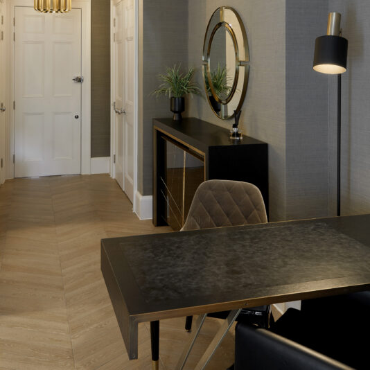 A modern interior space featuring the Modern Industrial Style Oak Veneer Desk in sleek black, accompanied by a brown cushioned chair. Against the wall stands a black console table topped with a round mirror and adorned with potted plants. The floor showcases a herringbone pattern, while a minimalistic white door adds to the modern industrial aesthetic in the background.