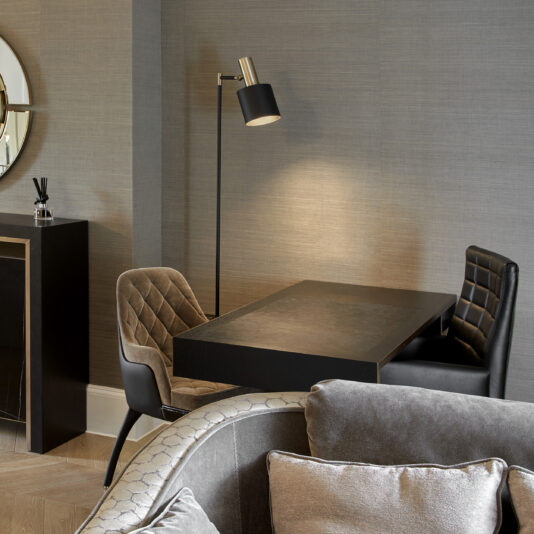 A cozy modern living room featuring a gray upholstered armchair in the foreground and a Modern Industrial Style Oak Veneer Desk with two cushioned chairs in the background. A sleek black floor lamp stands beside the desk, which complements the modern industrial style theme, while a round mirror decorates the gray wall.