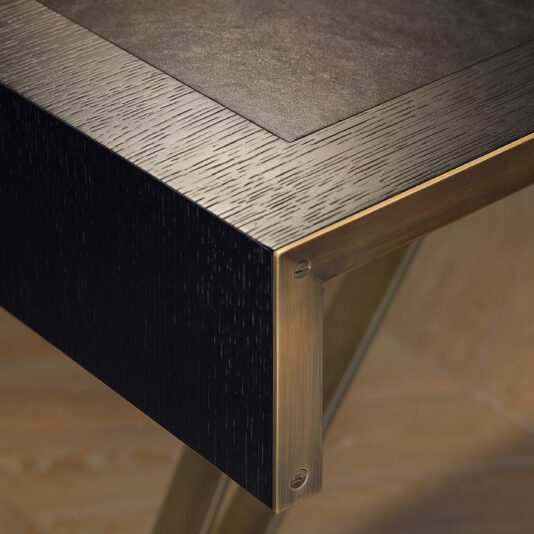 A close-up shot of a sleek, modern industrial style table corner highlights the sophisticated design of the Modern Industrial Style Oak Veneer Desk. The tabletop, with its dark wood appearance and textured surface, is elegantly framed by a metallic, brass-colored trim. The backdrop showcases a wooden floor that complements the oak veneer desk setting.