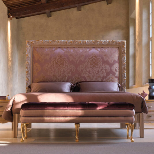 Luxurious bedroom with the Ornate Classic Style Bed With Tall Headboard, featuring an upholstered headboard adorned with intricate patterns. The bed is dressed in silky brown bedding and pillows. A cushioned bench with gold accents stands at the foot of the bed. Soft, natural light filters through a nearby window.