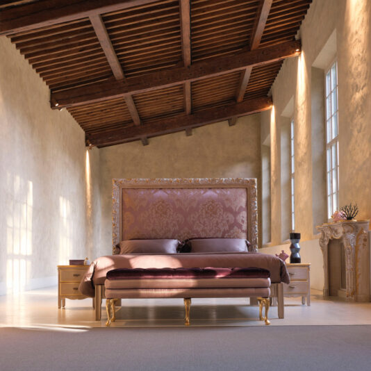 A luxurious bedroom features the Ornate Classic Style Bed With Tall Headboard, flanked by two matching nightstands. Natural light streams through tall windows, illuminating the wooden beamed ceiling and textured walls. A decorative console table stands to the right.