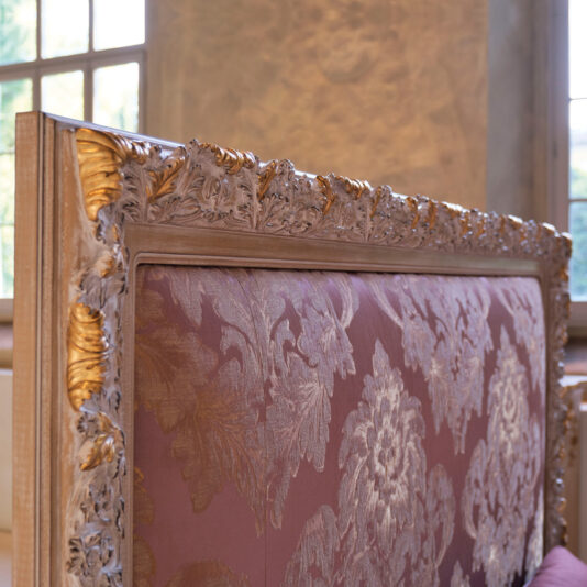 Close-up of the ornate top edge of a vintage chair. The wooden frame, reminiscent of an Ornate Classic Style Bed With Tall Headboard, is detailed with intricate carvings highlighted in gold paint. The upholstery features a floral damask pattern in shades of purple and gold. The background reveals a tall window and soft lighting.