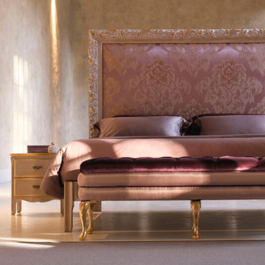 A luxurious bedroom features the Ornate Classic Style Bed With Tall Headboard, complete with a tall, gold-trimmed headboard upholstered in pink fabric adorned with an intricate pattern. The bed is complemented by matching pillows and a burgundy bench at its foot. A wooden nightstand with a drawer sits beside the bed.