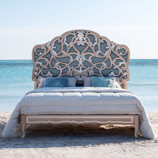 An Ornate Rococo Style Bed With Tall Headboard, featuring a majestic and intricately carved design, is set on a sandy beach near the ocean. The bed is neatly made with light-colored bedding and multiple pillows, creating a striking contrast against the clear blue sky and turquoise waters in the background.