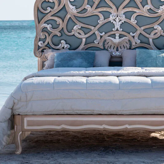 Ornate Rococo Style Bed With Tall Headboard The Ornate Rococo Style Bed with Tall Headboard sits on a beach with turquoise waters in the background. The bedding is in light shades, and several blue and gray decorative pillows are arranged against the headboard. The bed frame features elegant carvings.