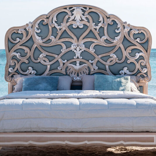 Ornate Rococo Style Bed With Tall Headboard An Ornate Rococo Style Bed With Tall Headboard, featuring intricate carvings and scrollwork, stands luxuriously on a beach facing the sea. The bed is adorned with white linens and several blue and grey pillows arranged against the tall headboard.