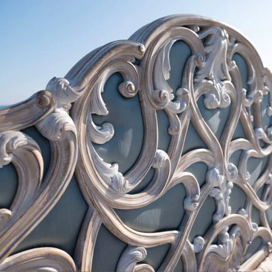 Ornate Rococo Style Bed With Tall Headboard Close-up of the Ornate Rococo Style Bed With Tall Headboard, highlighting its intricate, swirling patterns in the tall, carved wood headboard. The light beige wood boasts an antiqued finish, set against a pastel blue background. This photo beautifully captures the craftsmanship of its elaborate design under a clear sky.